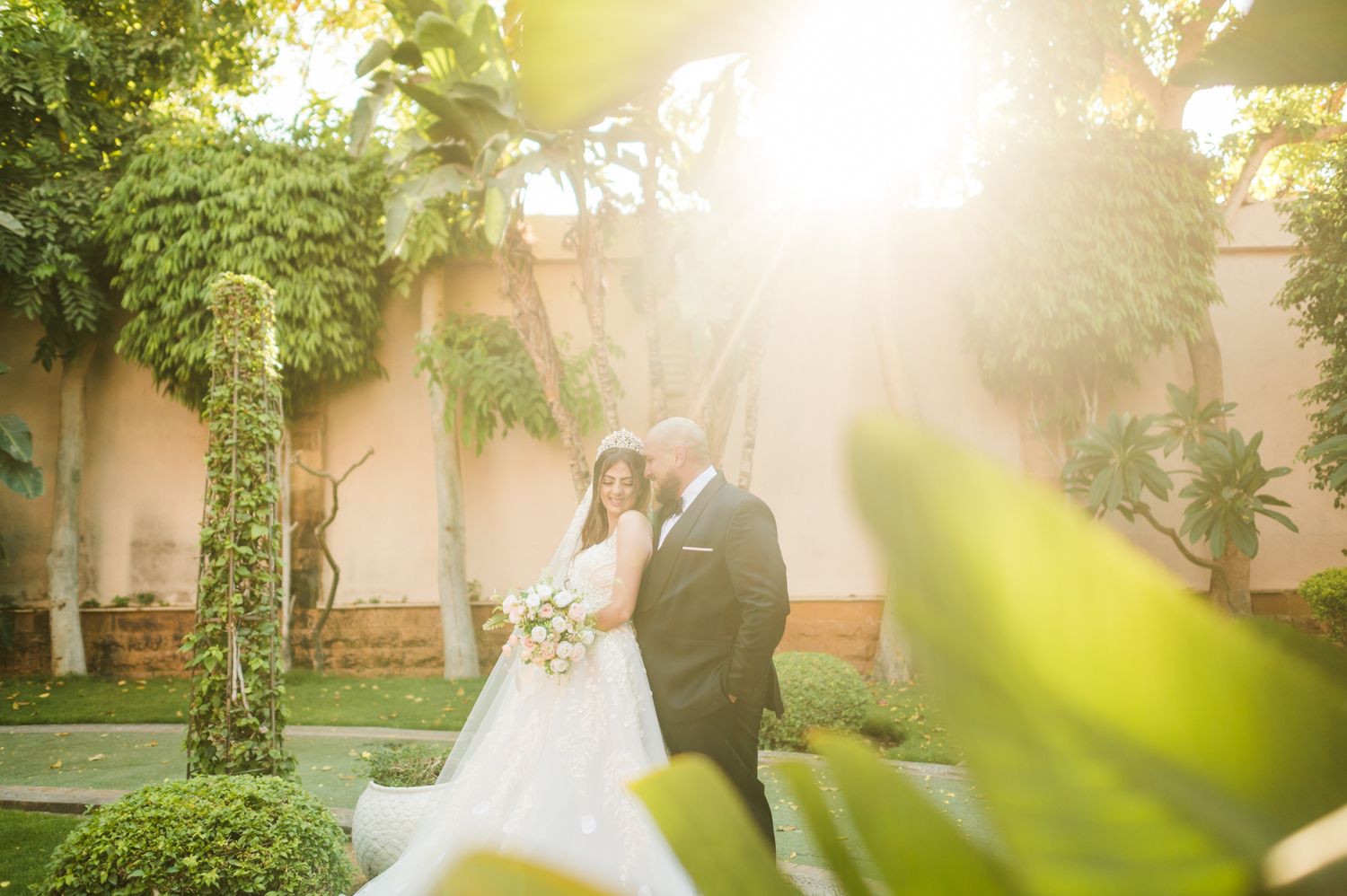 A wedding couple embraces in a sunlit garden with climbing vines and palm trees.