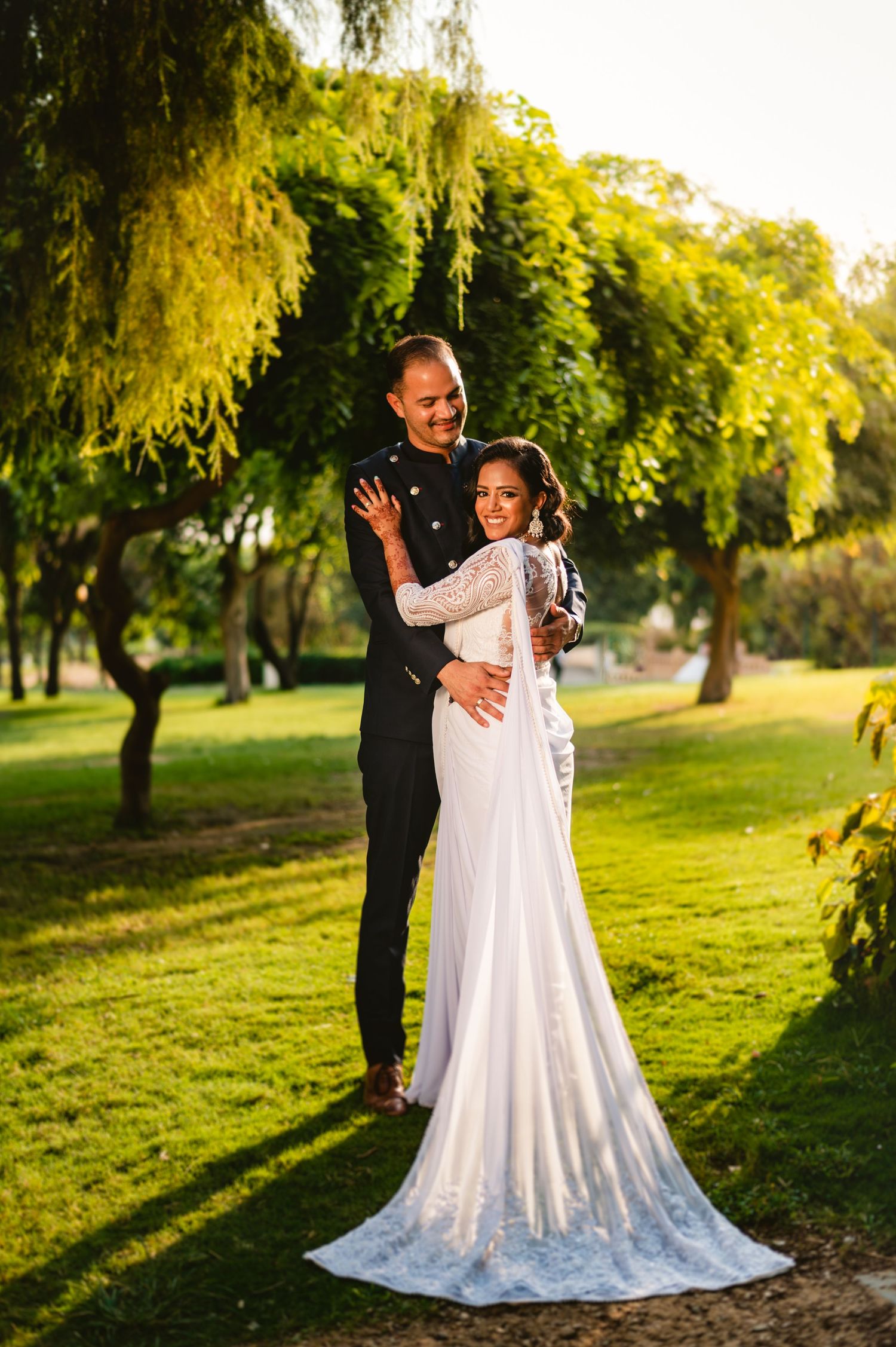 A newlywed couple embraces in a lush garden setting with a long white wedding gown trailing behind.