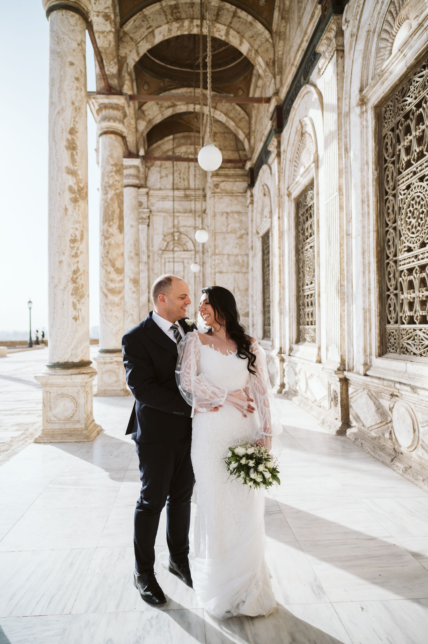 A couple poses in an elegant stone colonnade with ornate architectural details.