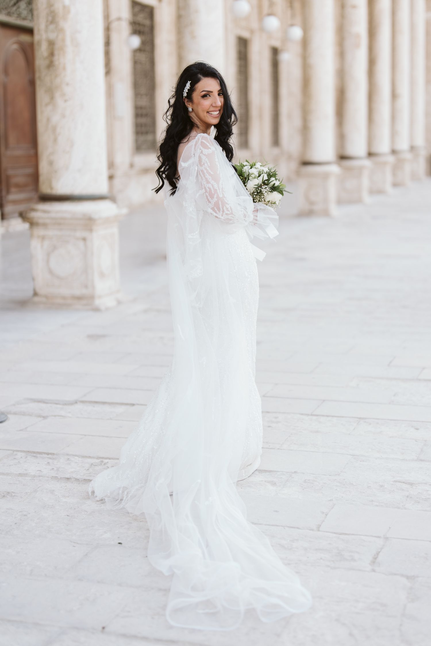 Bride in long-sleeved lace wedding gown poses in front of classic columned architecture.
