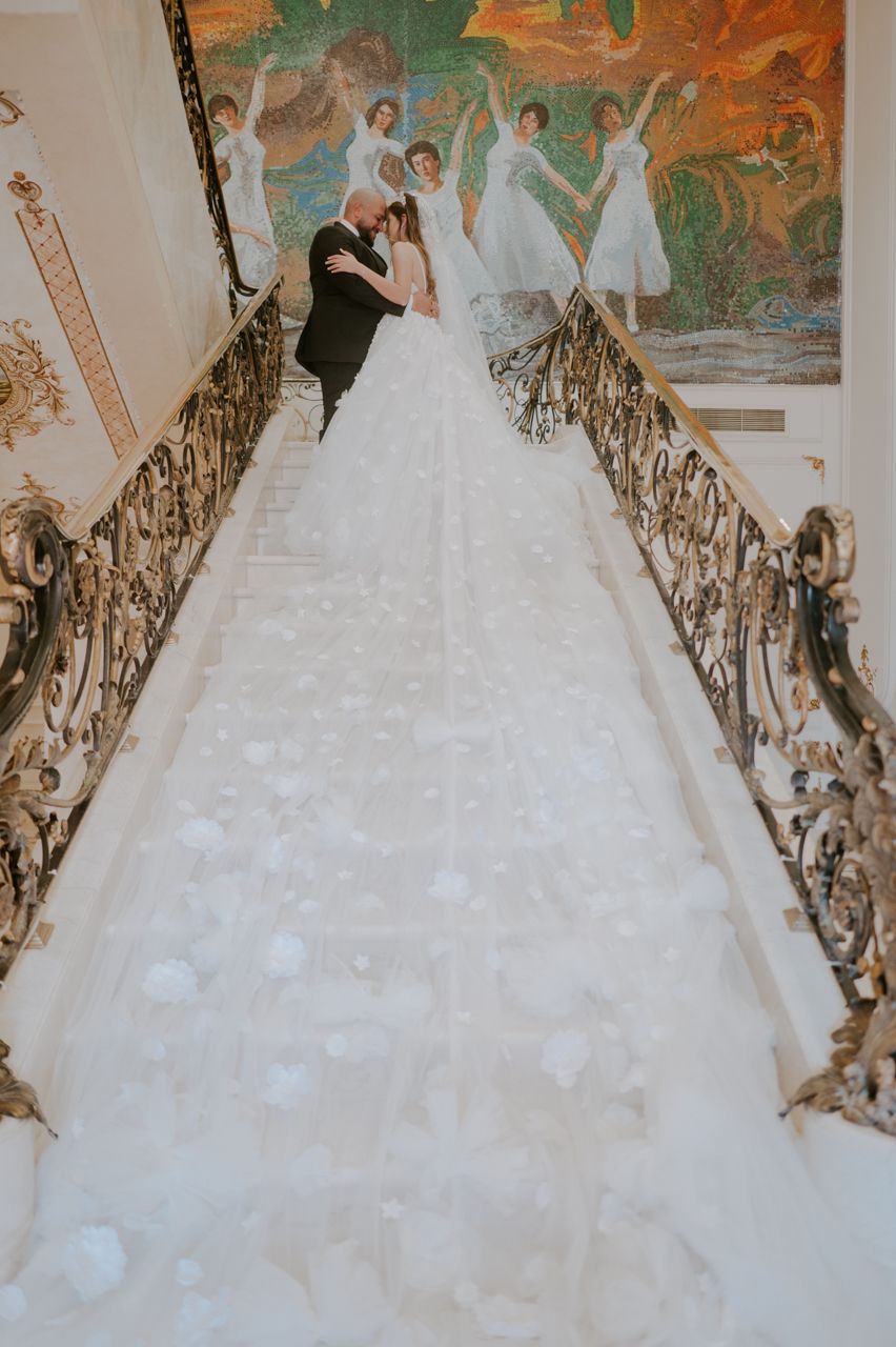 A couple stands on ornate grand staircase with detailed murals and gilded railings.