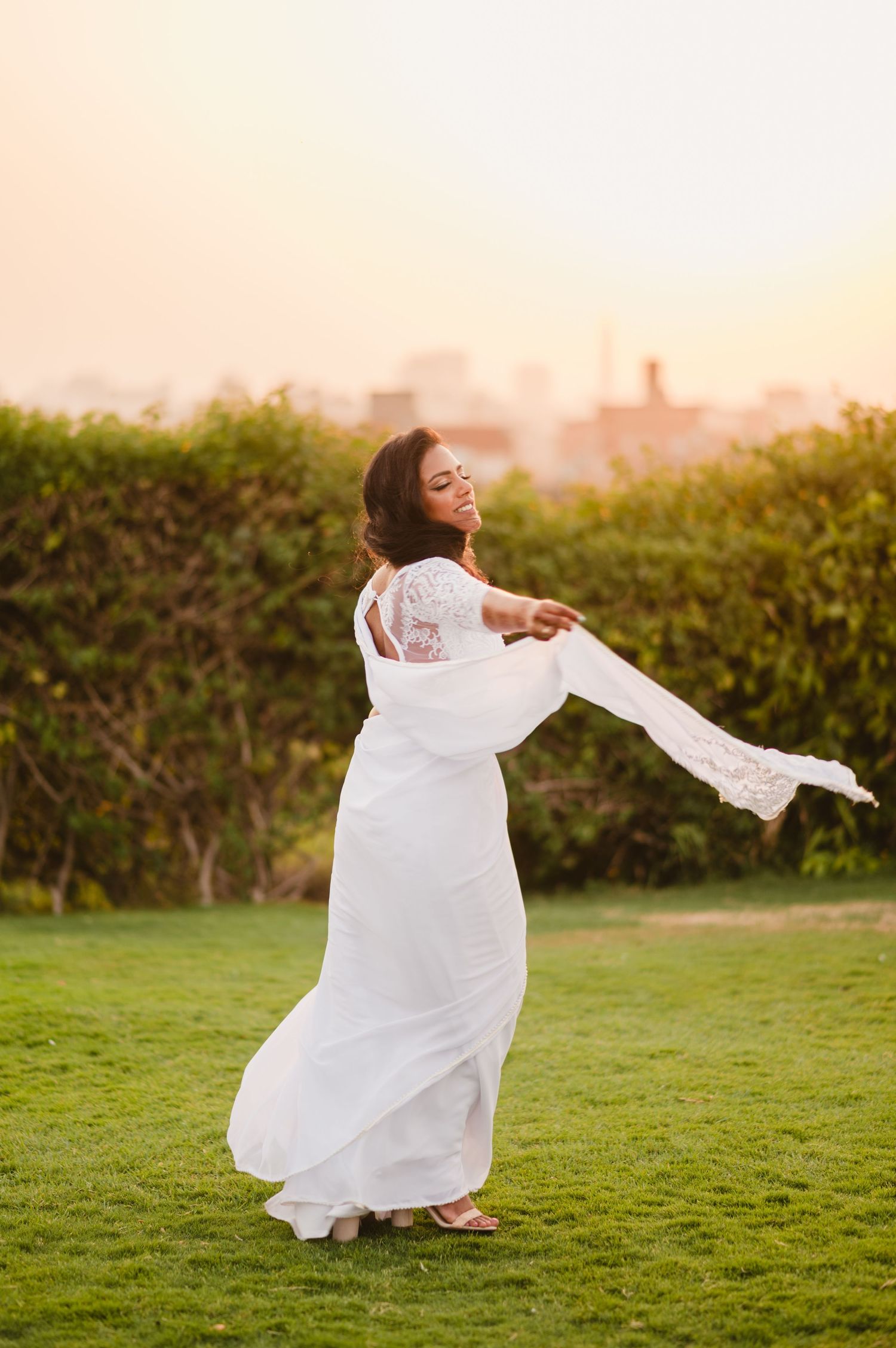 A bride in a flowing white dress twirls joyfully on a grassy lawn during golden hour.