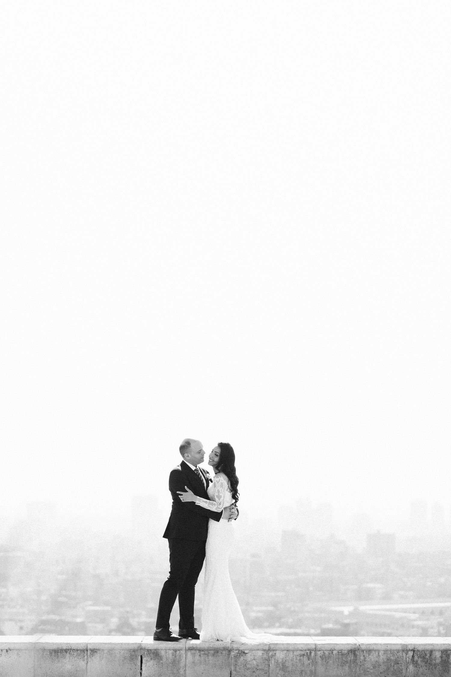 A romantic black and white wedding photo of a couple embracing on a rooftop overlooking a city skyline.