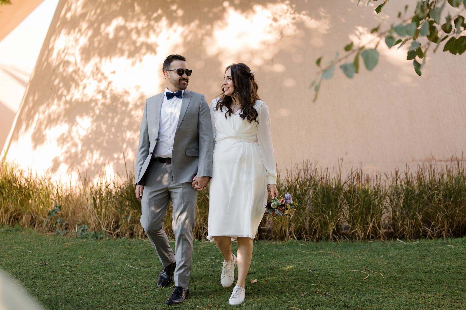 Elegantly dressed couple strolling together on grass against a sunlit wall.