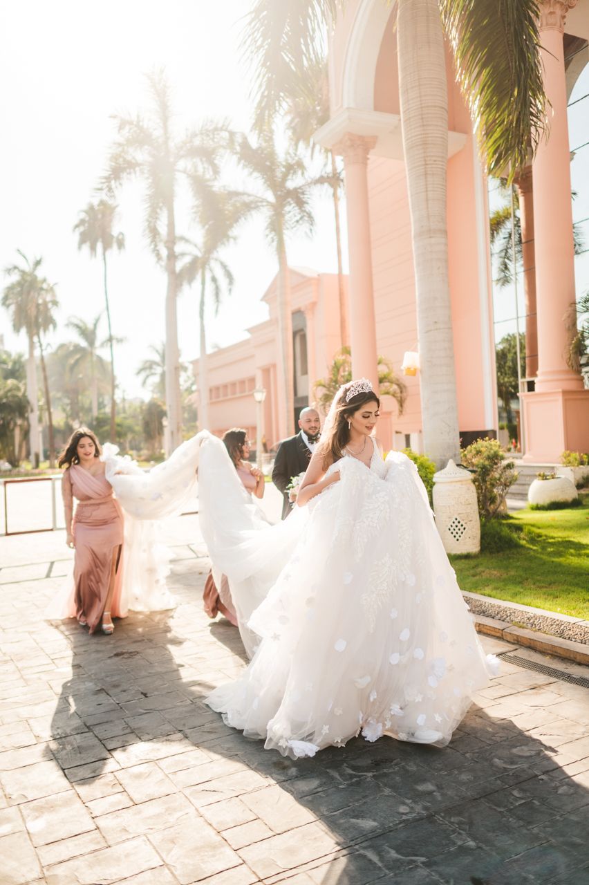 A bride in a flowing white gown poses with attendants against a pink building with palm trees.