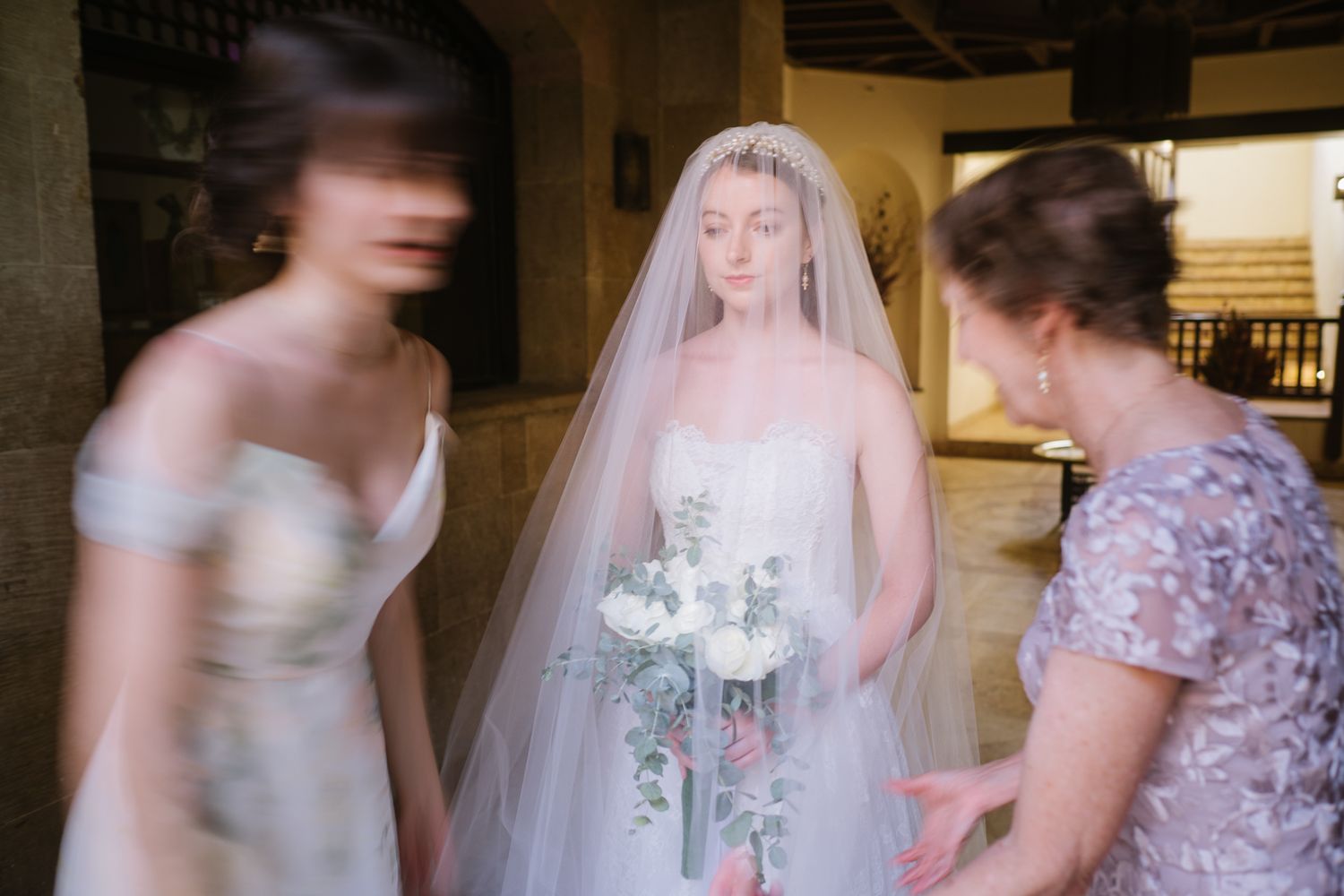 A bride in a white wedding dress and veil holds a bouquet while preparing with attendants.