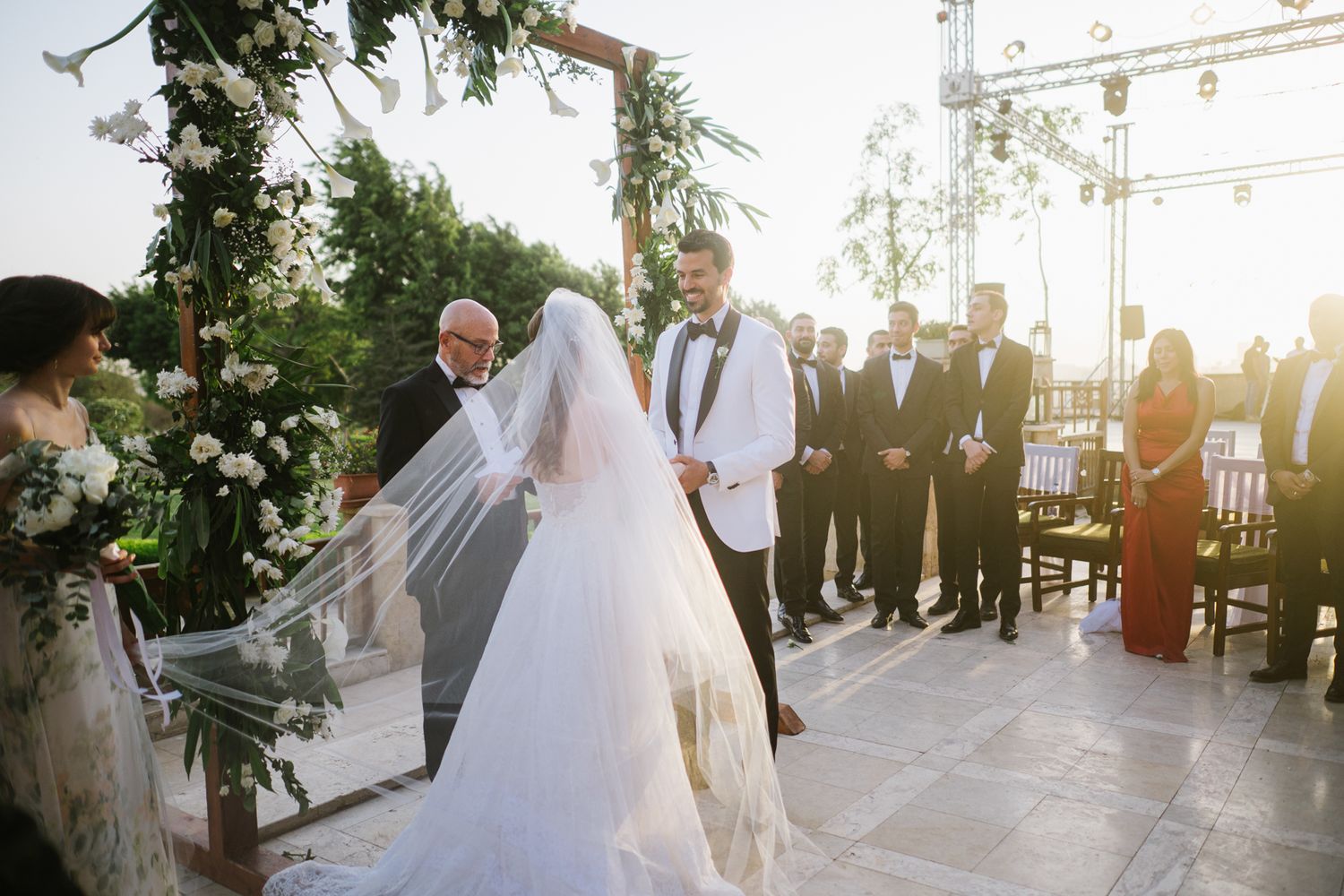 A wedding ceremony taking place outdoors during golden hour under a floral arch with guests gathered.