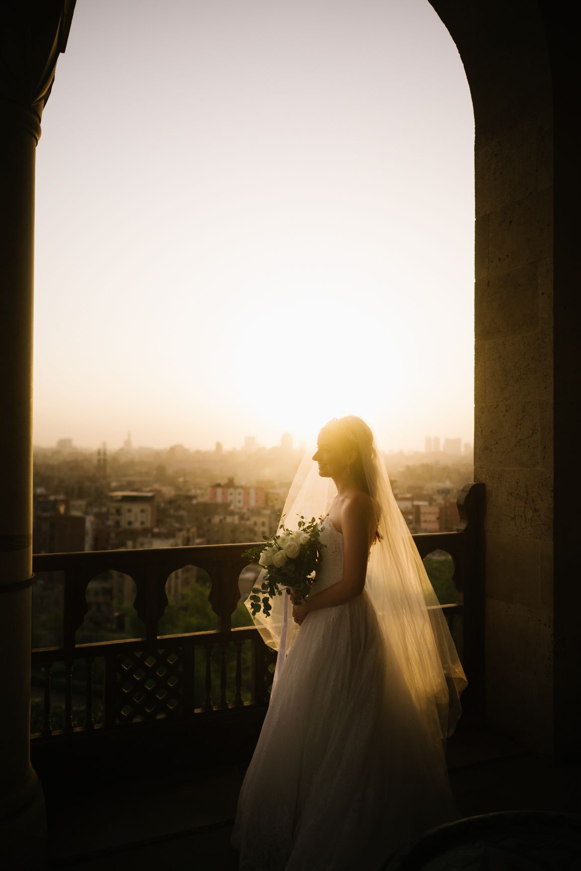 A silhouetted bride in flowing dress gazes at sunset through an arched doorway.