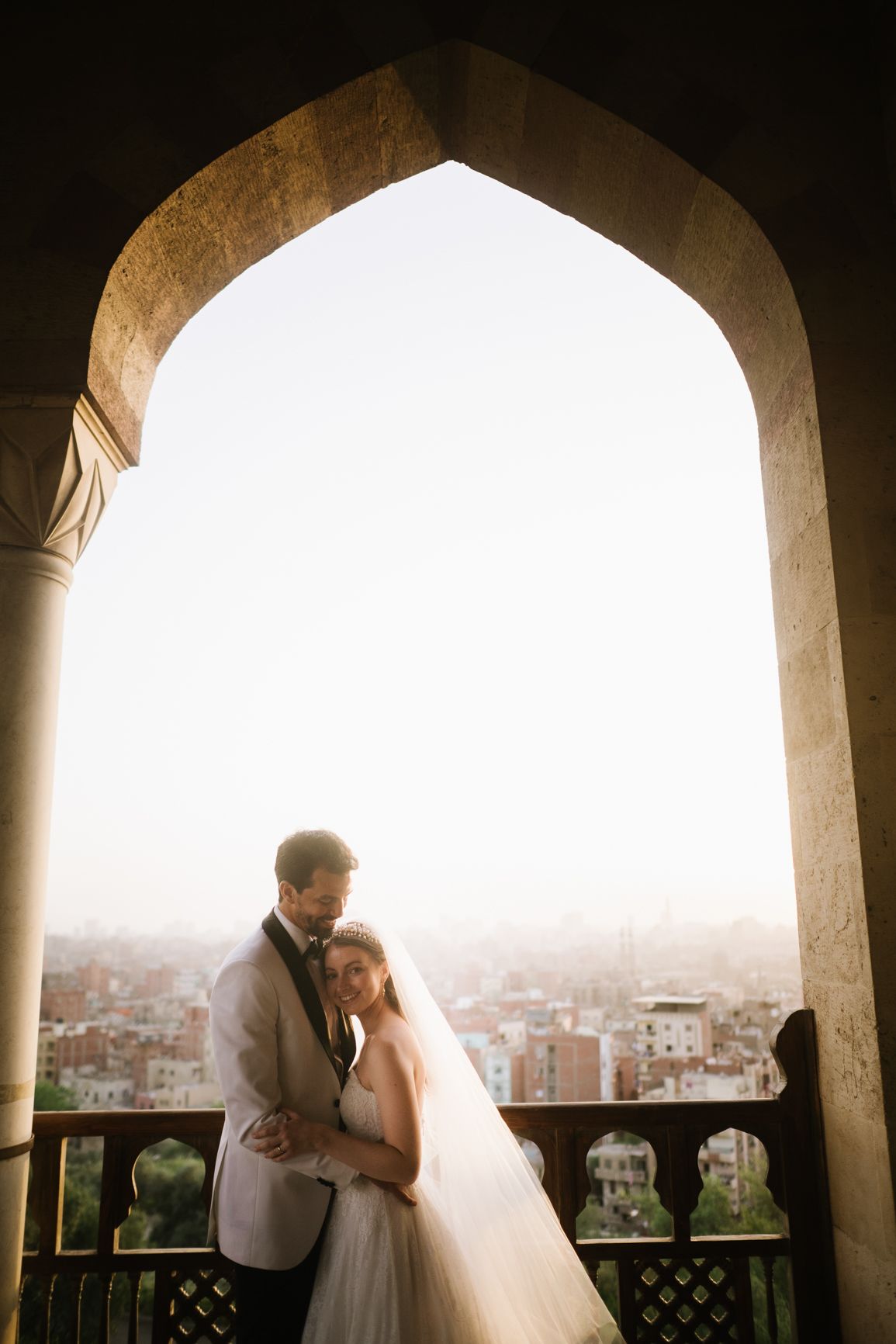 A romantic wedding portrait framed by a grand archway overlooking a cityscape.