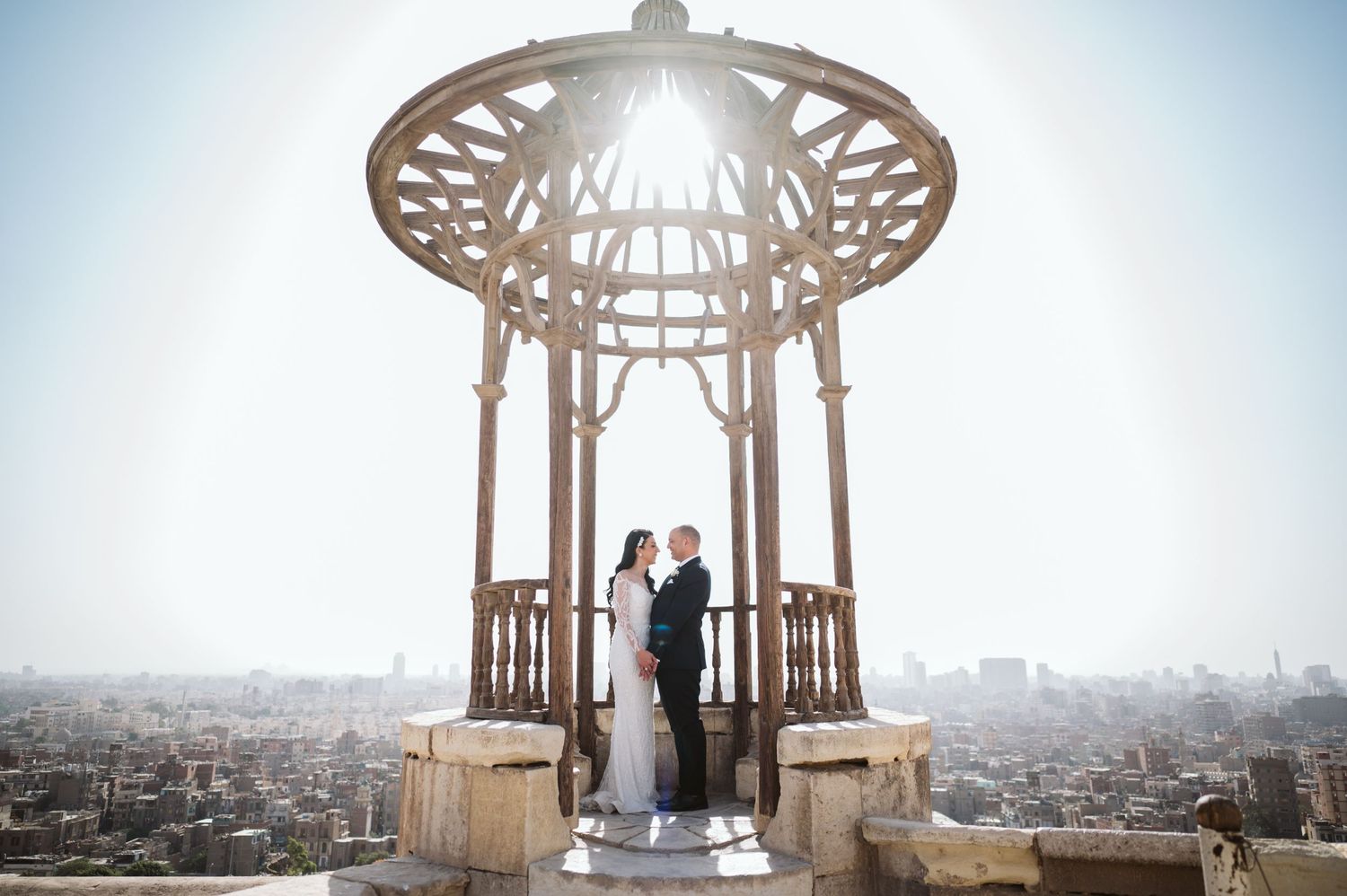 A newlywed couple stands in an ornate gazebo overlooking a cityscape.