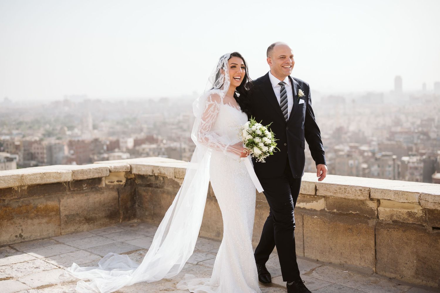 A couple walks together on a stone terrace with a stunning city backdrop.