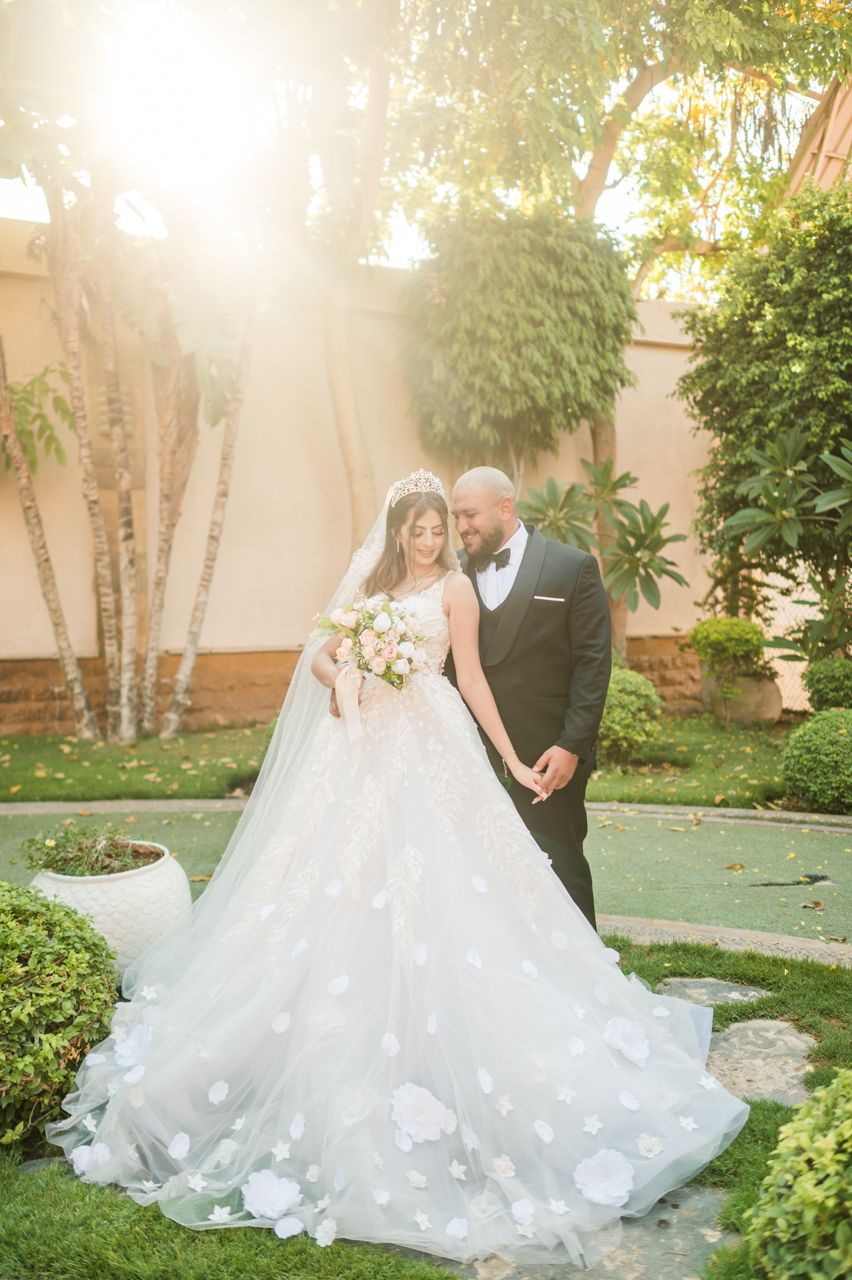 A couple poses in formal wedding attire in a lush garden setting with warm sunlight.