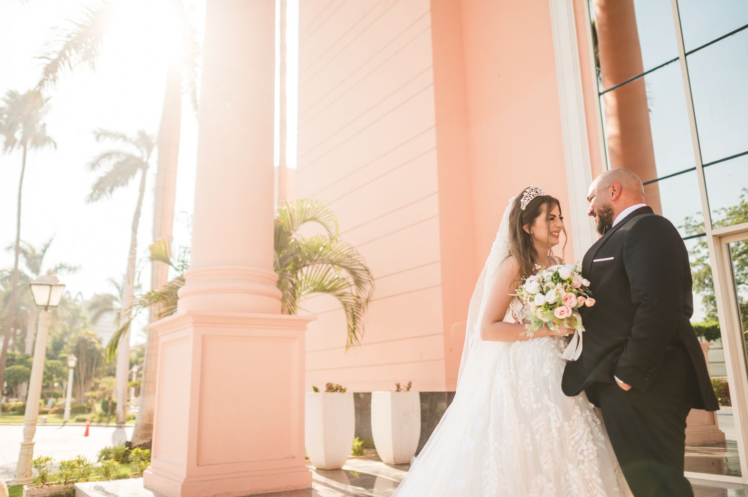 Wedding couple shares tender moment in front of pink building with palm trees at sunset.