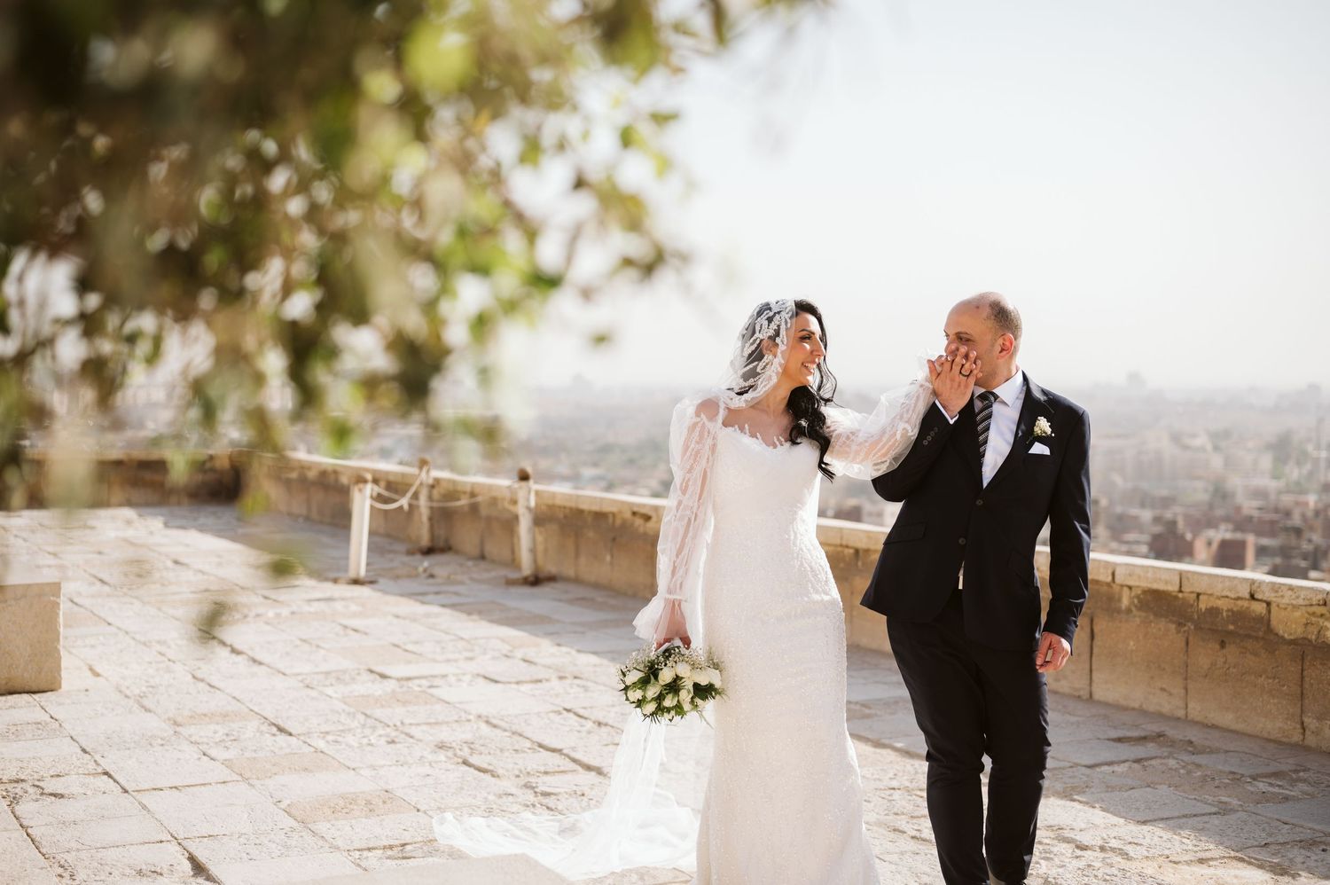Newlyweds share a candid moment while walking along a sunlit stone terrace.