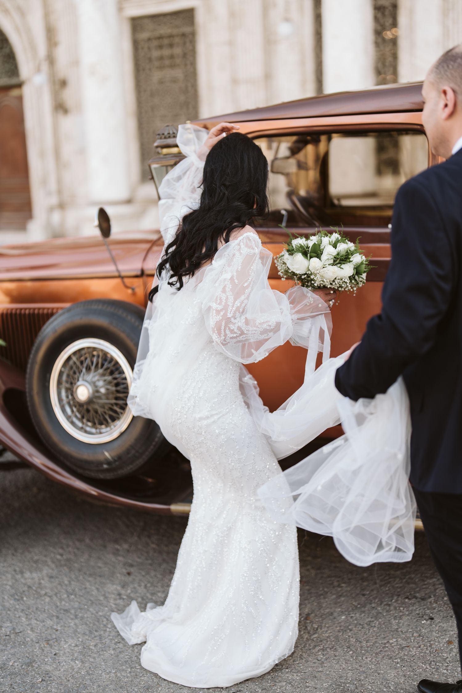 Bride in flowing white dress stands by vintage car with classic architectural backdrop.