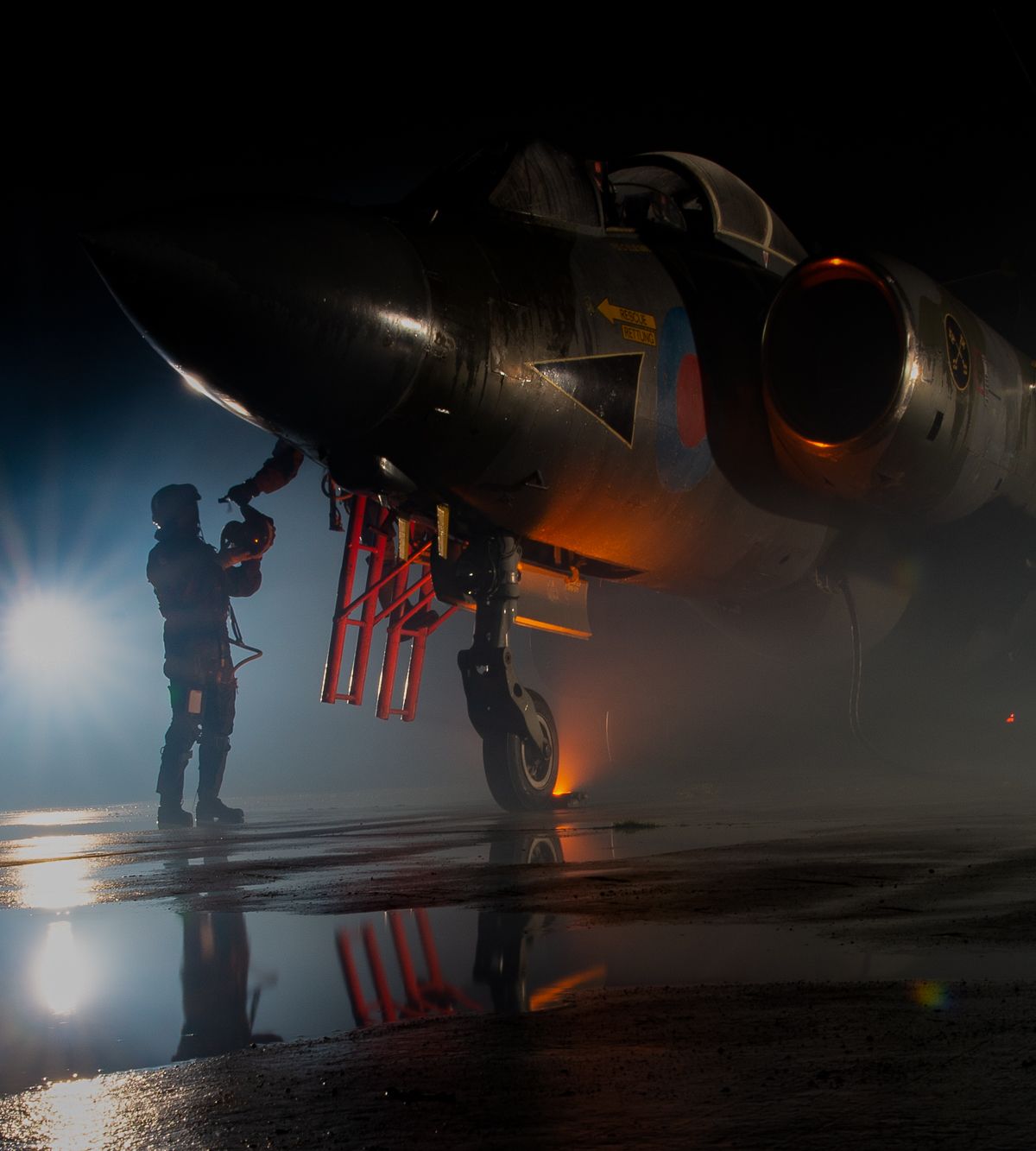 Silhouettes of ground crew working on a military aircraft at night with dramatic lighting and fog.