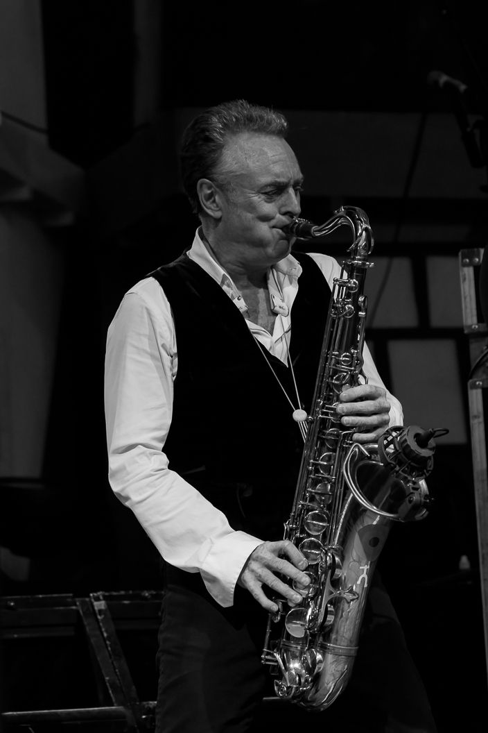 Musician performing on saxophone in black and white wearing formal attire on stage.