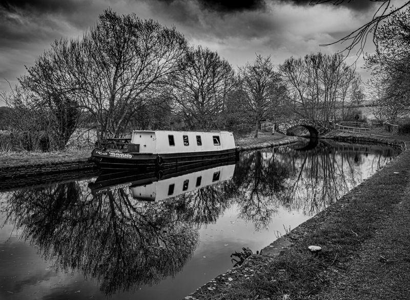 Black and white photograph of canal boats reflecting in still water with bare trees in winter.