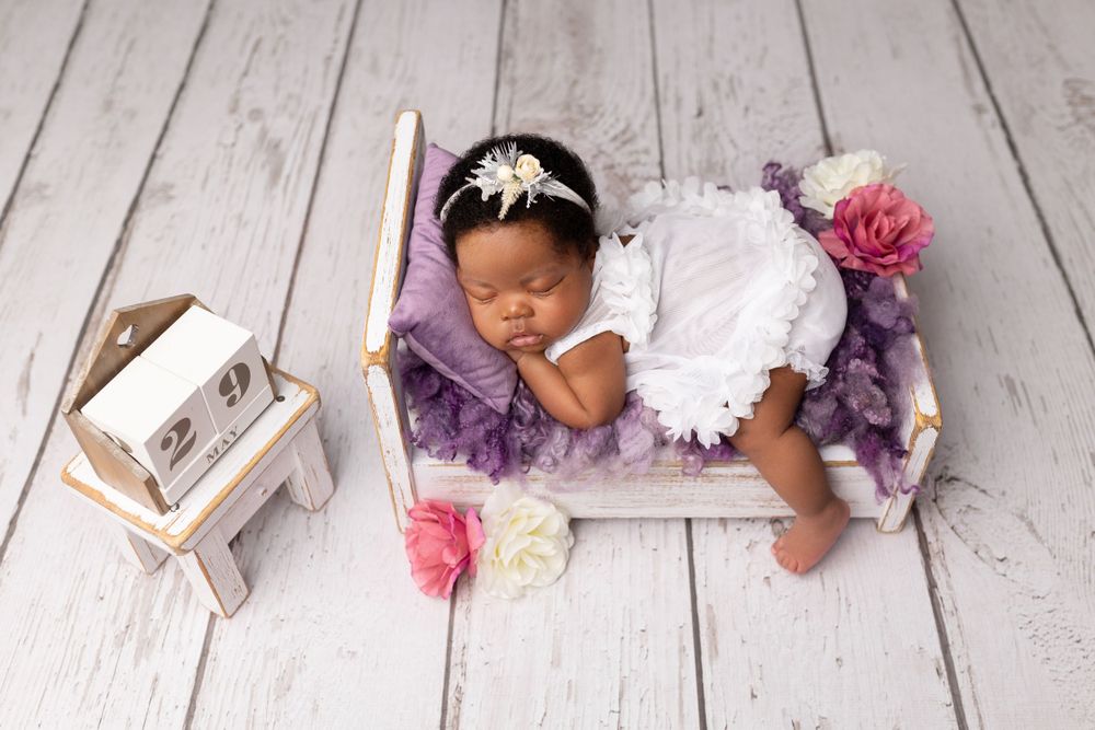 Sleeping baby in white dress on purple cushion with pink flowers and decorative frame.