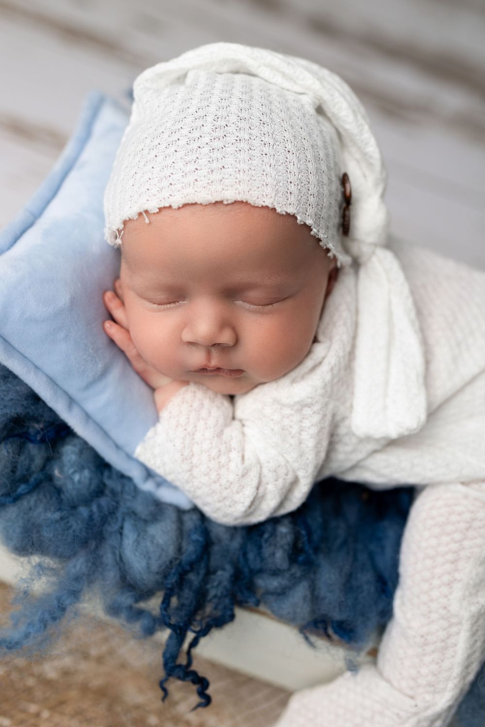 Sleeping newborn wrapped in white knit bonnet and blanket on blue cushion during professional photo session.