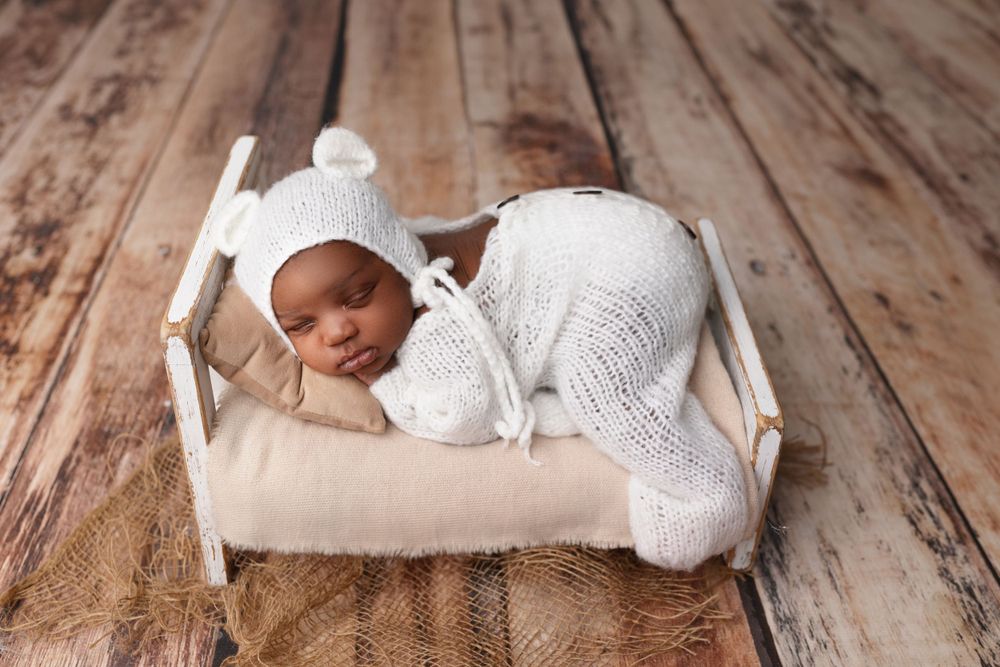 Newborn in white knit outfit sleeping on miniature wooden bed.