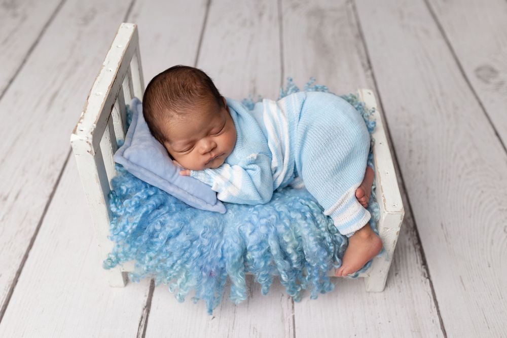 Newborn sleeping in small white bed with blue tulle skirt on wooden floor during photo shoot.