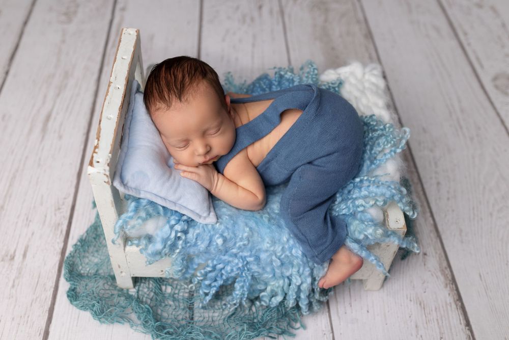 Peaceful sleeping infant in blue outfit on blue lace cushion in white crib during photo session.