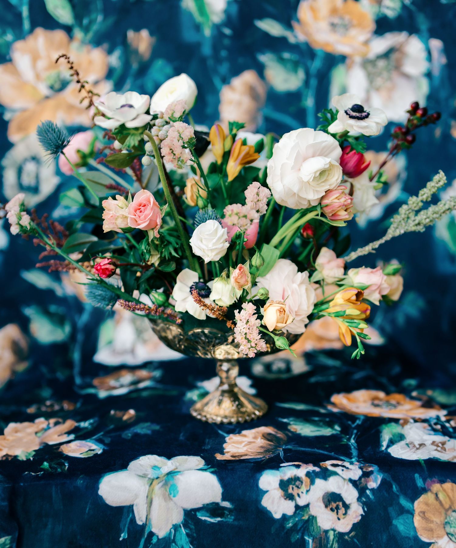 Luxurious floral centerpiece with roses and mixed blooms in a gold vessel against blue background.