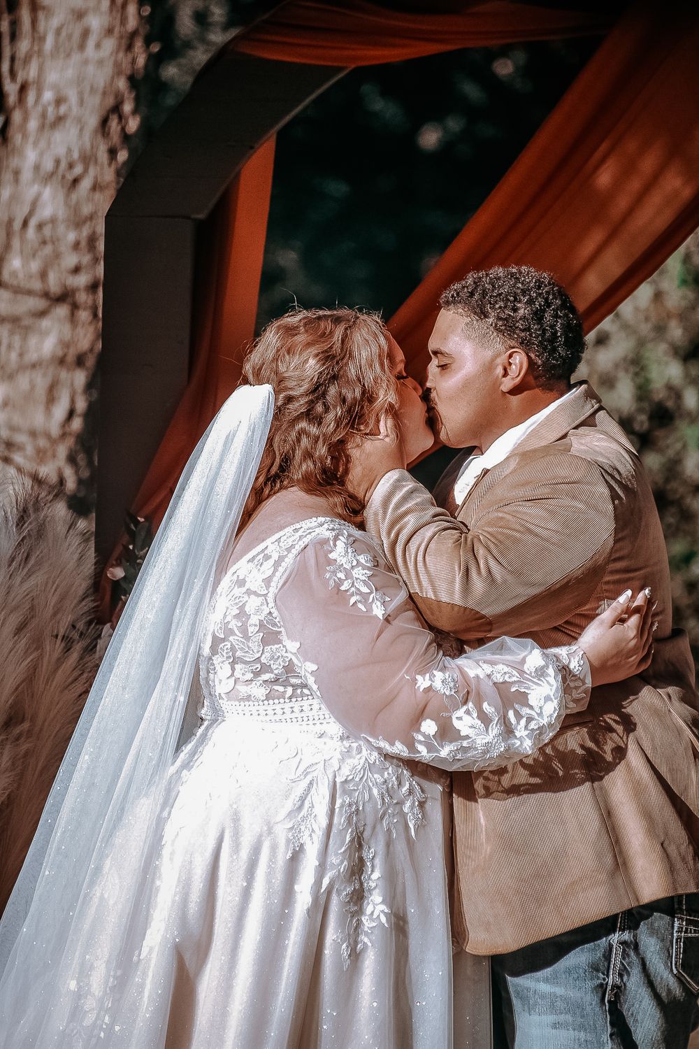 A romantic first kiss as newlyweds under a wooden arch decorated with orange fabric.