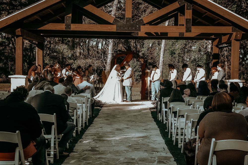 A wedding ceremony takes place in a rustic covered pavilion filled with guests seated in white chairs.