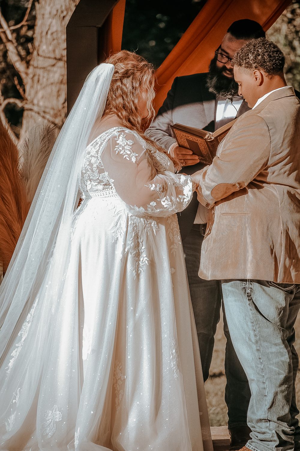 Side view of wedding ceremony showing detailed lace dress and casual groom attire during ring exchange.