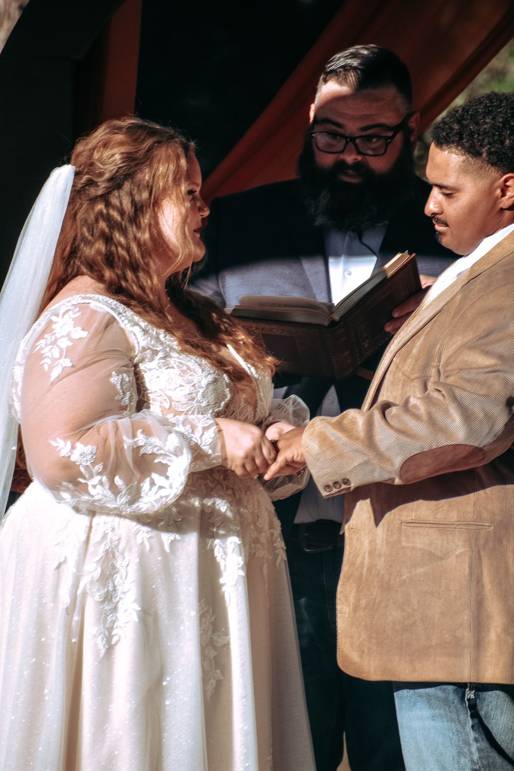 Close-up of wedding ceremony showing ring exchange with detailed lace dress in sunlight.