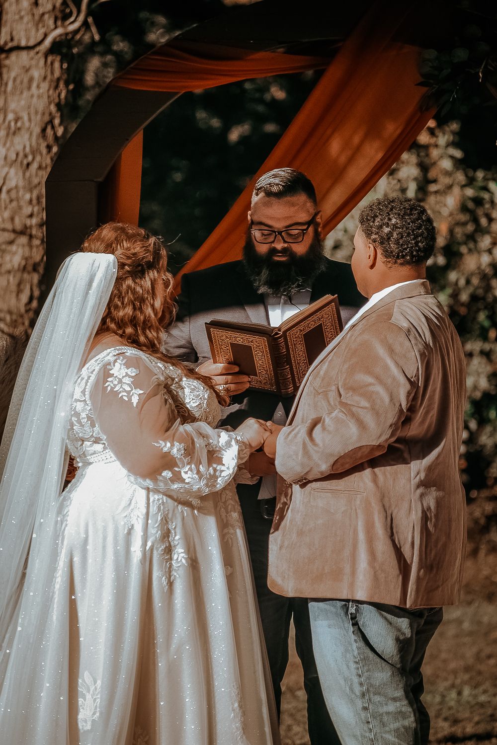 A couple exchanges vows during an outdoor wedding ceremony under a wooden arch with orange draping.