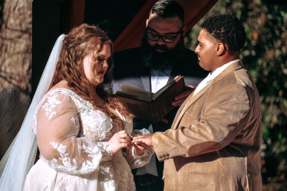 Emotional ring exchange moment during outdoor wedding ceremony with wooden arch backdrop.