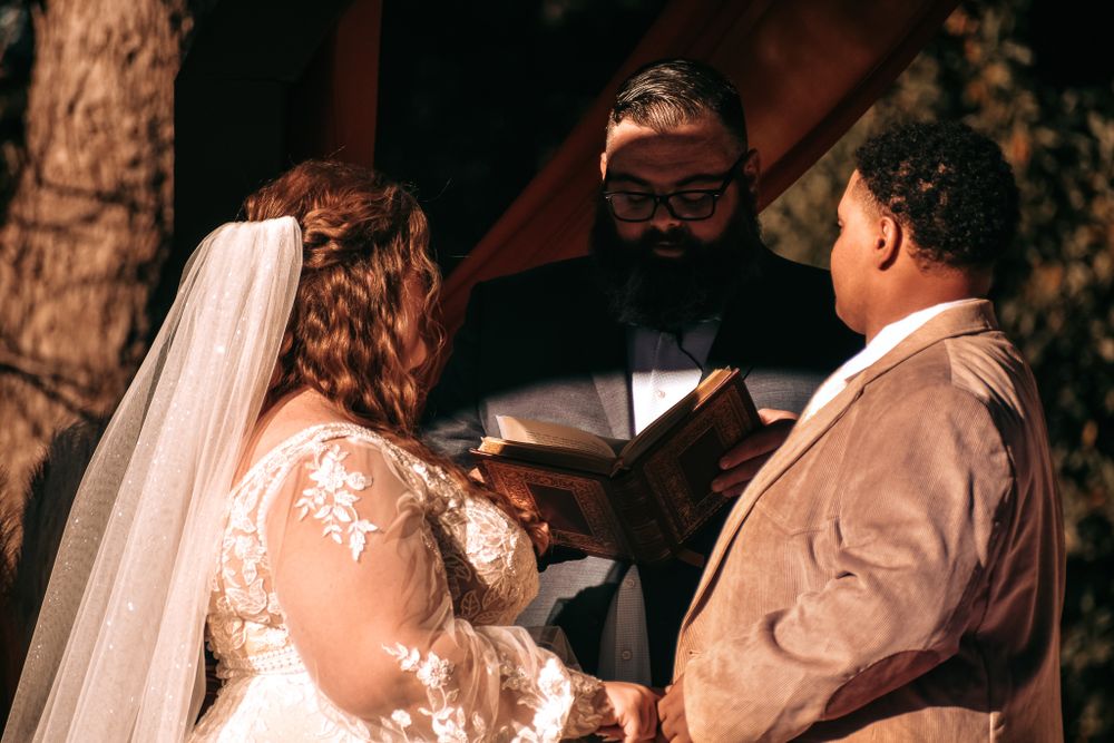Intimate wedding ceremony moment under wooden arbor with officiant reading from book.