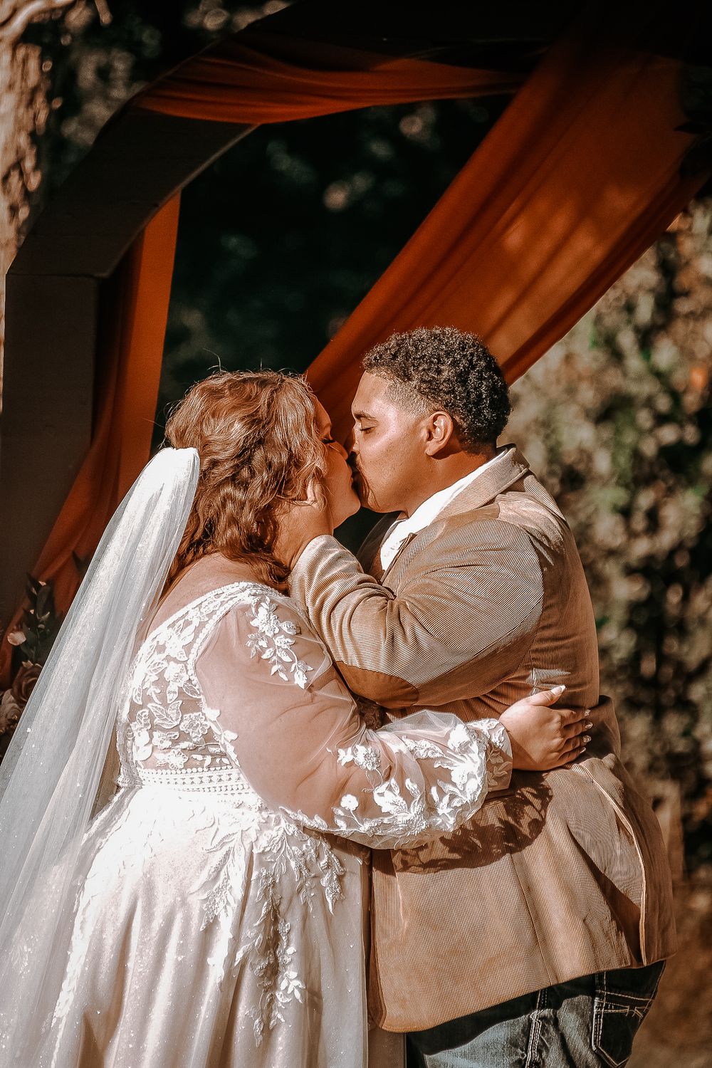 Newlyweds share an intimate moment during their wedding ceremony beneath an orange-draped arch.