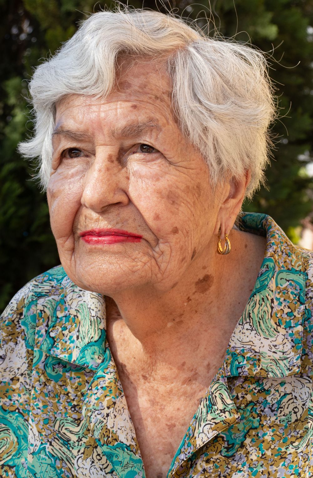Senior in turquoise floral blouse poses outdoors with natural greenery background.