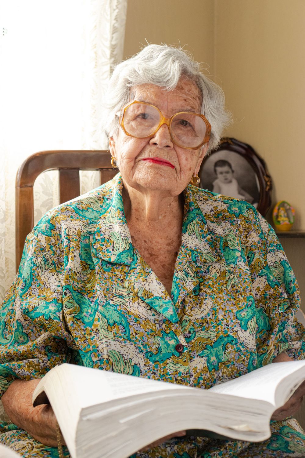 Elderly reader in glasses and floral dress examines pages of an open book.