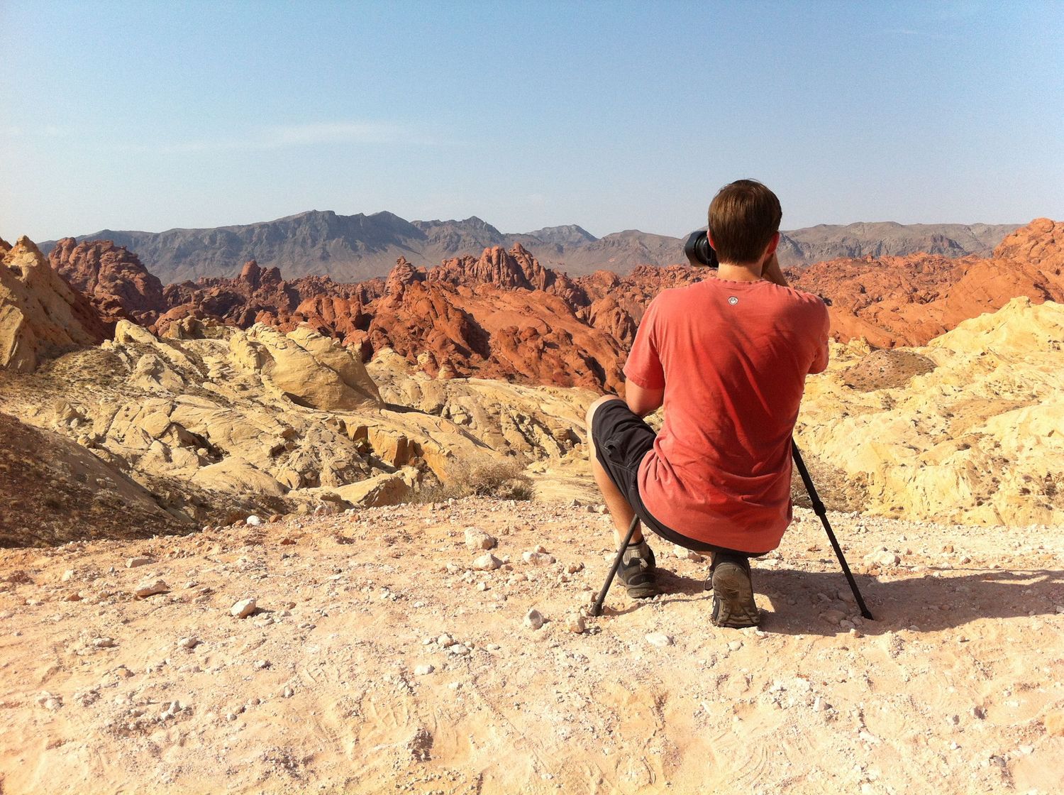 Nature photographer, Alex Kent crouches behind tripod facing toward tan, red and grey rock layer mountain ranges.