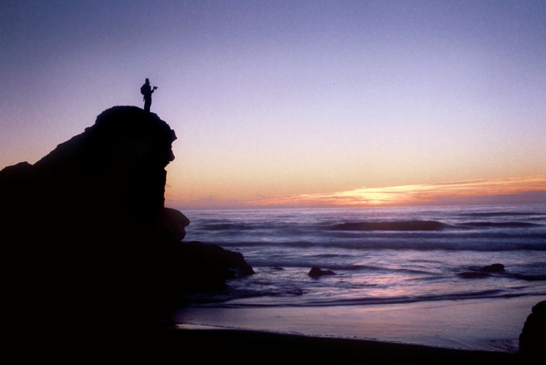 Arkansas nature photographer, Alex Kent stands on top of massive rock along the Pacific coast beach with sunset behind