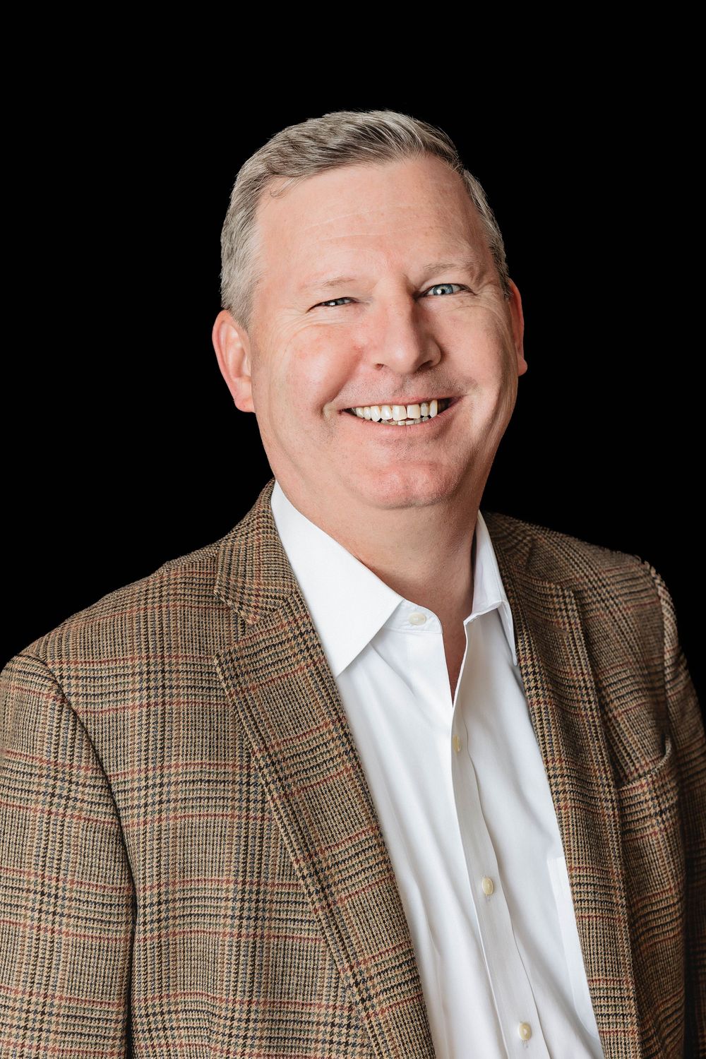 Professional headshot of a smiling person wearing a brown plaid blazer and white shirt against a black background.
