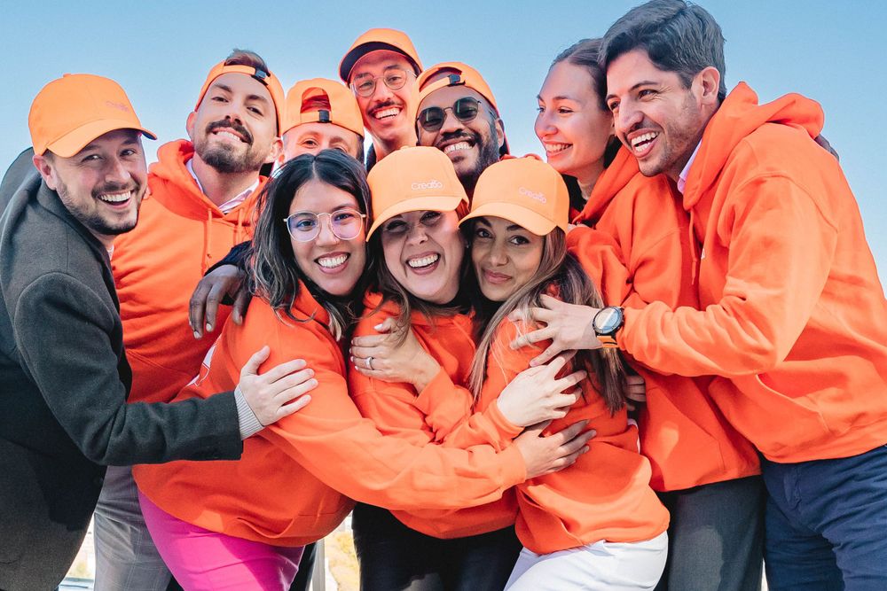 Group of friends in bright orange hoodies laughing and hugging outdoors.