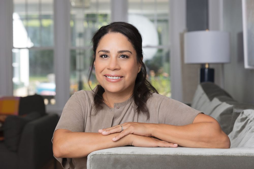 Professional headshot in home office setting with gray couch and lamp.