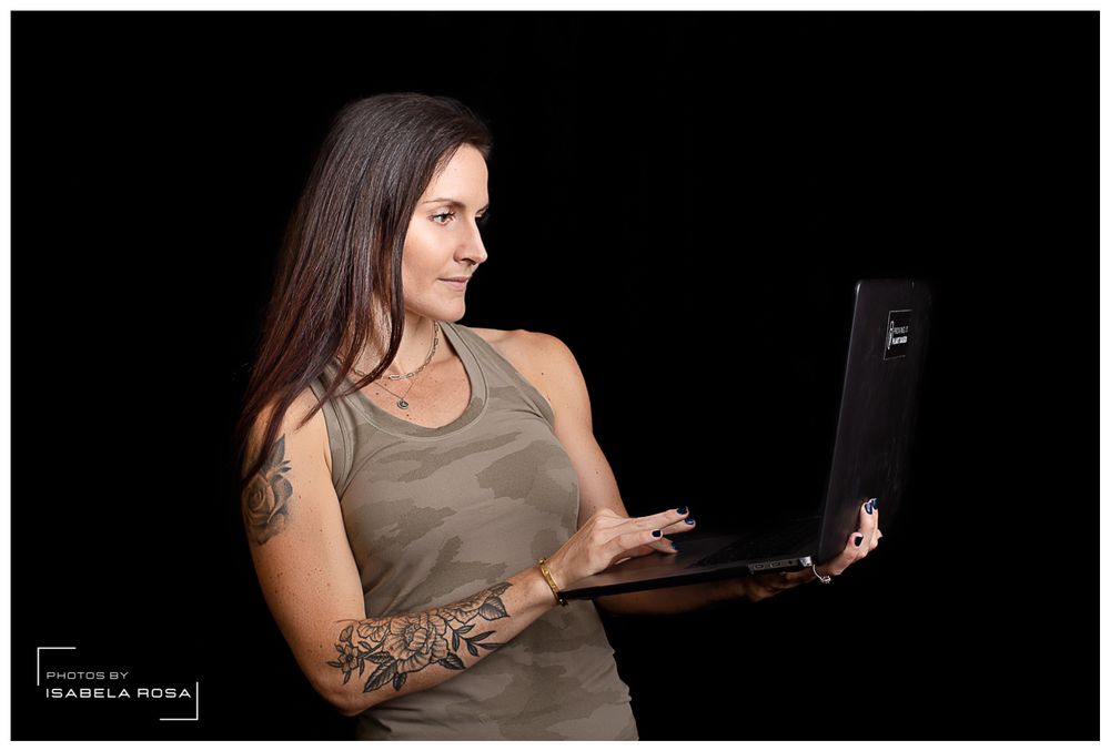 Woman in tank top with arm tattoos using laptop computer against black background.