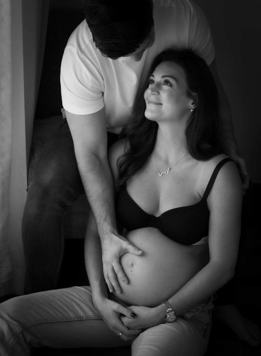 Pregnant woman in jeans and bra sat looking at partner whose hand is on her pregnant belly - maternity photoshoot