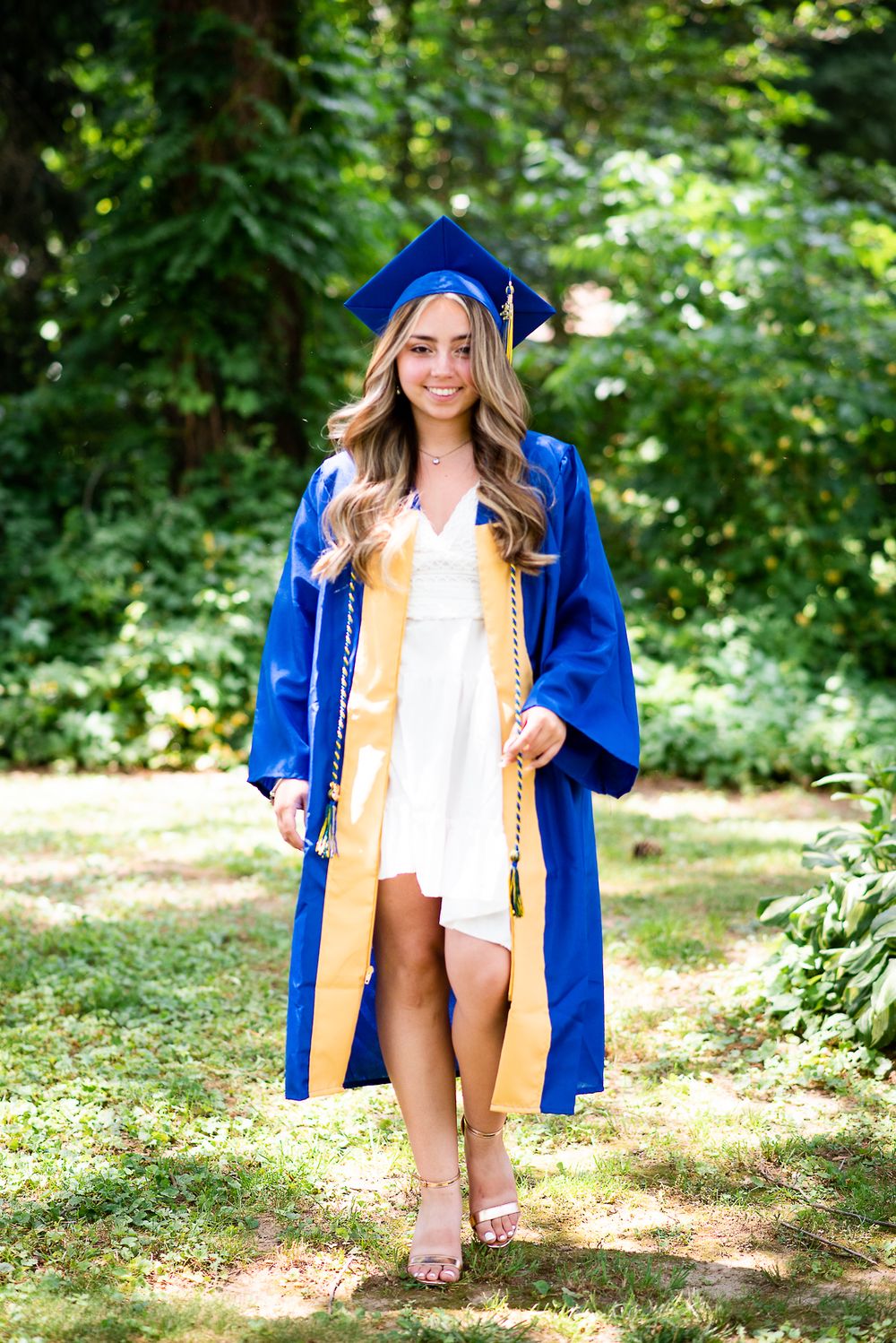 Graduate in blue cap and gown poses in a sunny garden setting wearing a white dress.