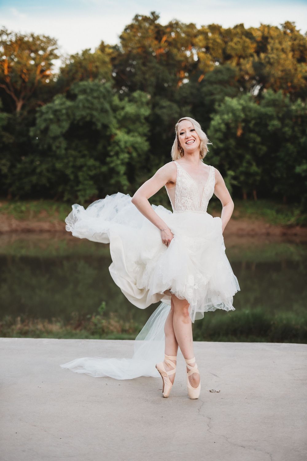 Ballet dancer in wedding dress strikes elegant pose en pointe by lakeside at sunset.