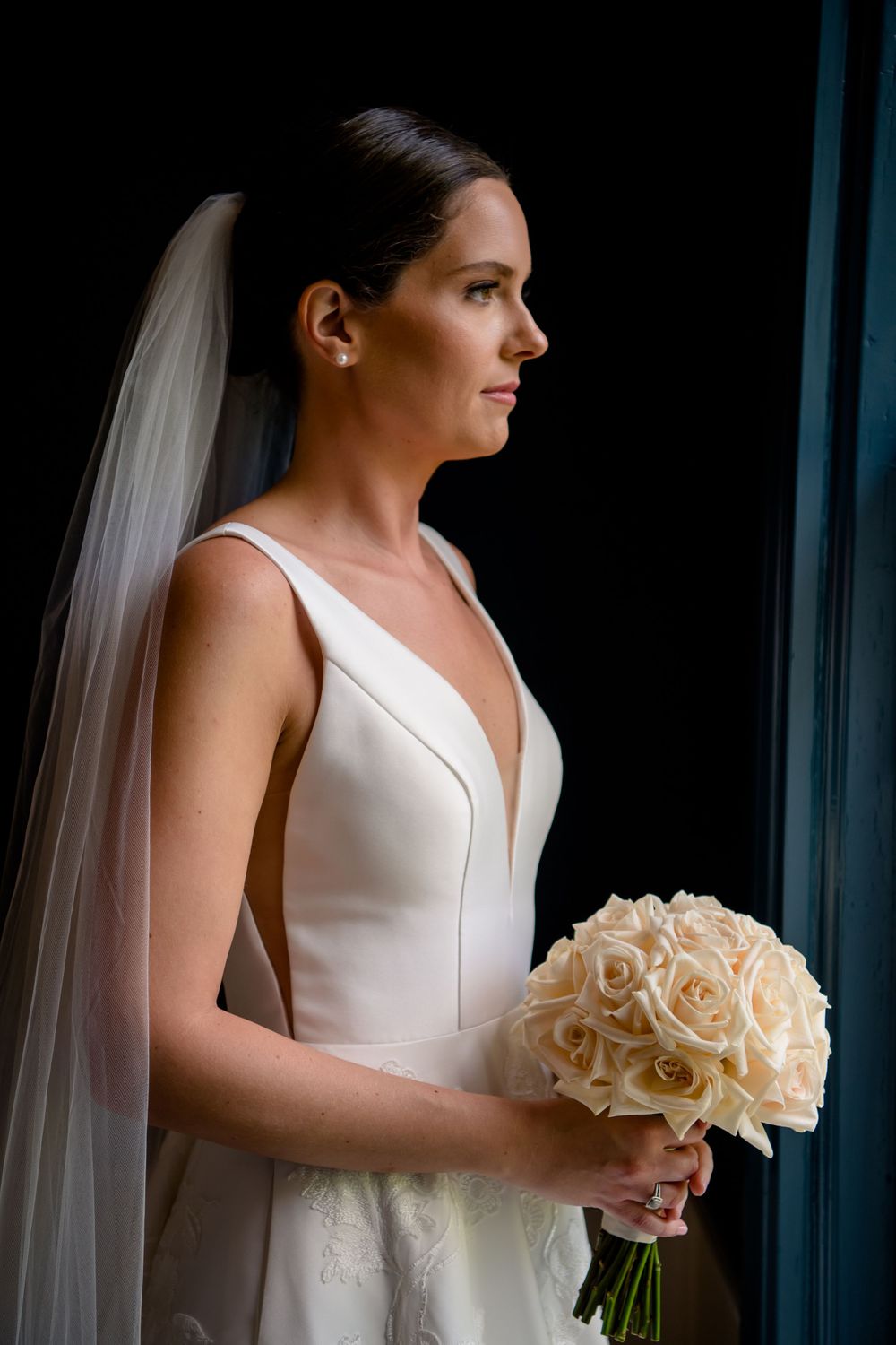 Dramatic side profile of bride in minimal white gown holding cream rose bouquet.