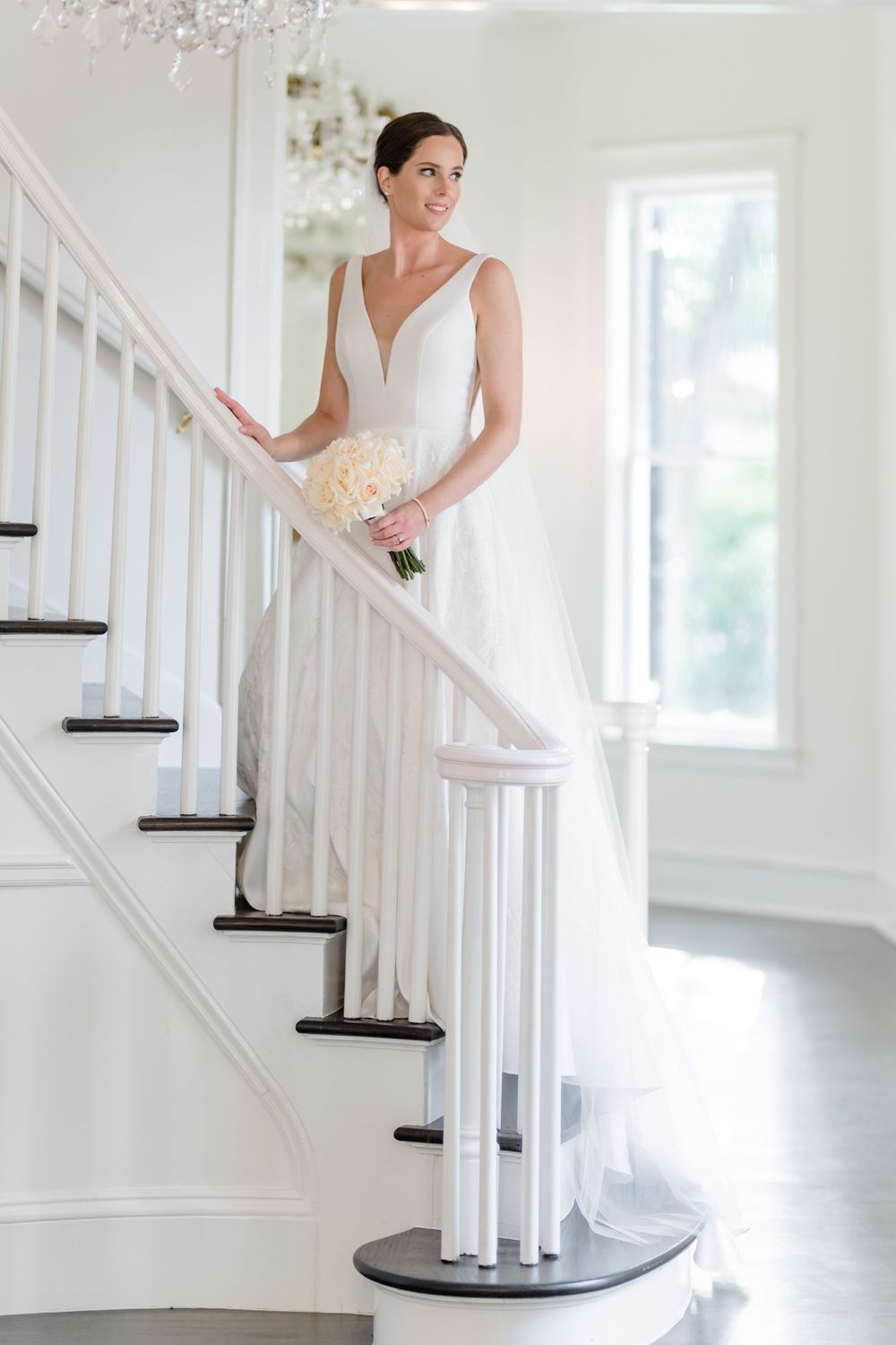 Elegant bridal portrait on white staircase holding cream roses in classic wedding gown.