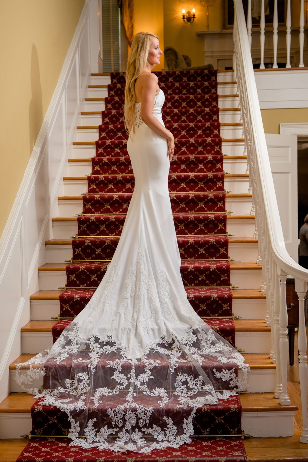 A wedding dress with lace train cascades down red carpeted stairs in an elegant interior.